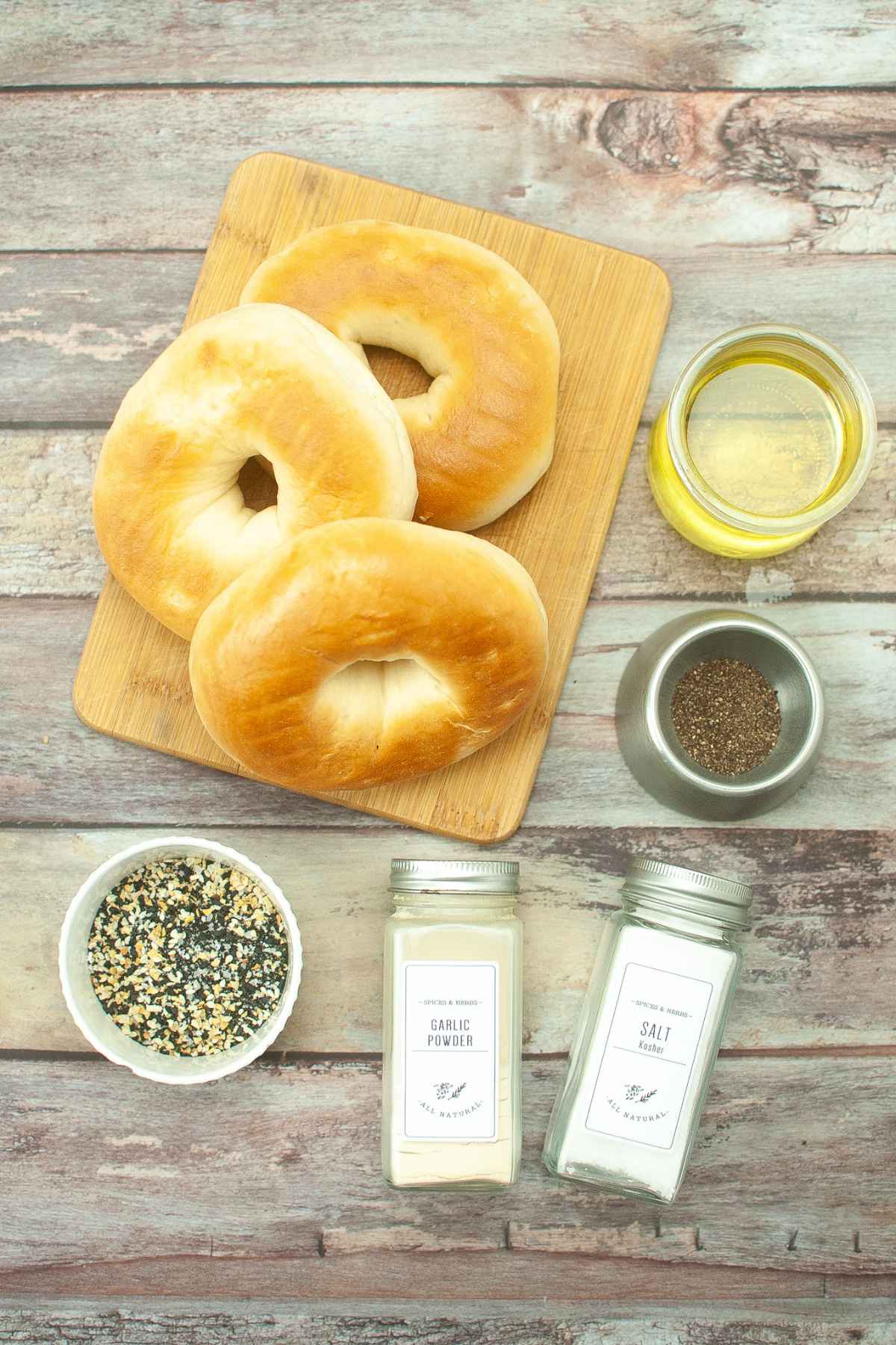 Three bagels on a cutting board with a jar of olive oil, black pepper, garlic powder, and everything seasoning.