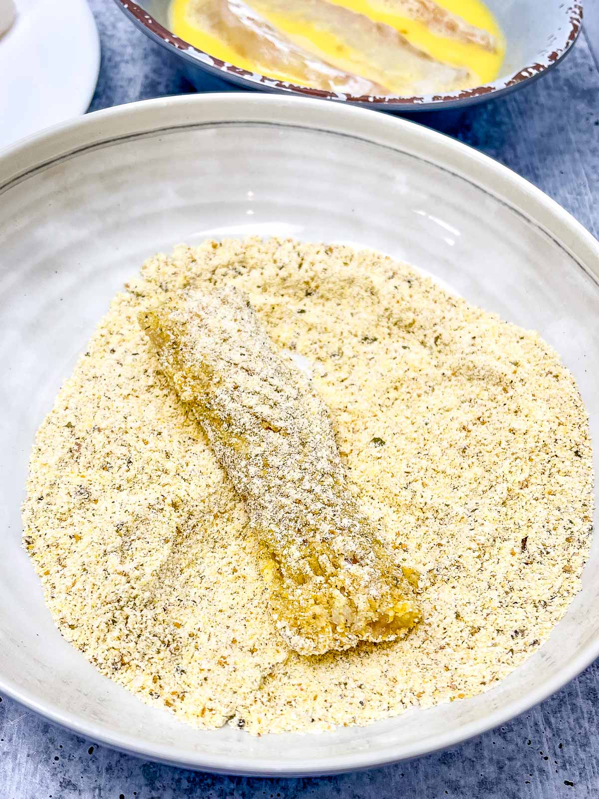 Cornmeal breading in a bowl with a fish stick inside the bowl coated with the corn flour.