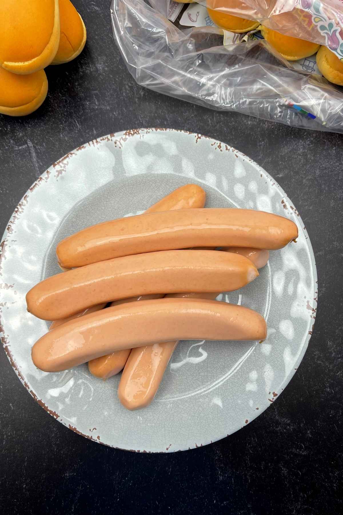 Raw hotdogs on a plate with hot dog rolls in the background.