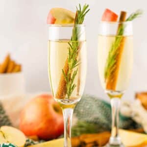 Featured image with two glasses of apple cider mimosa garnished with apple slice, cinnamon stick, and rosemary.