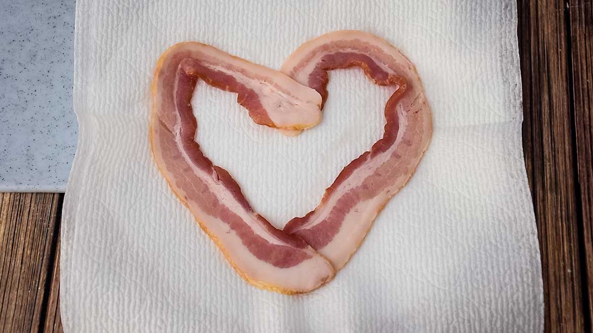 Raw bacon in a heart shape on a paper towel lined plate.