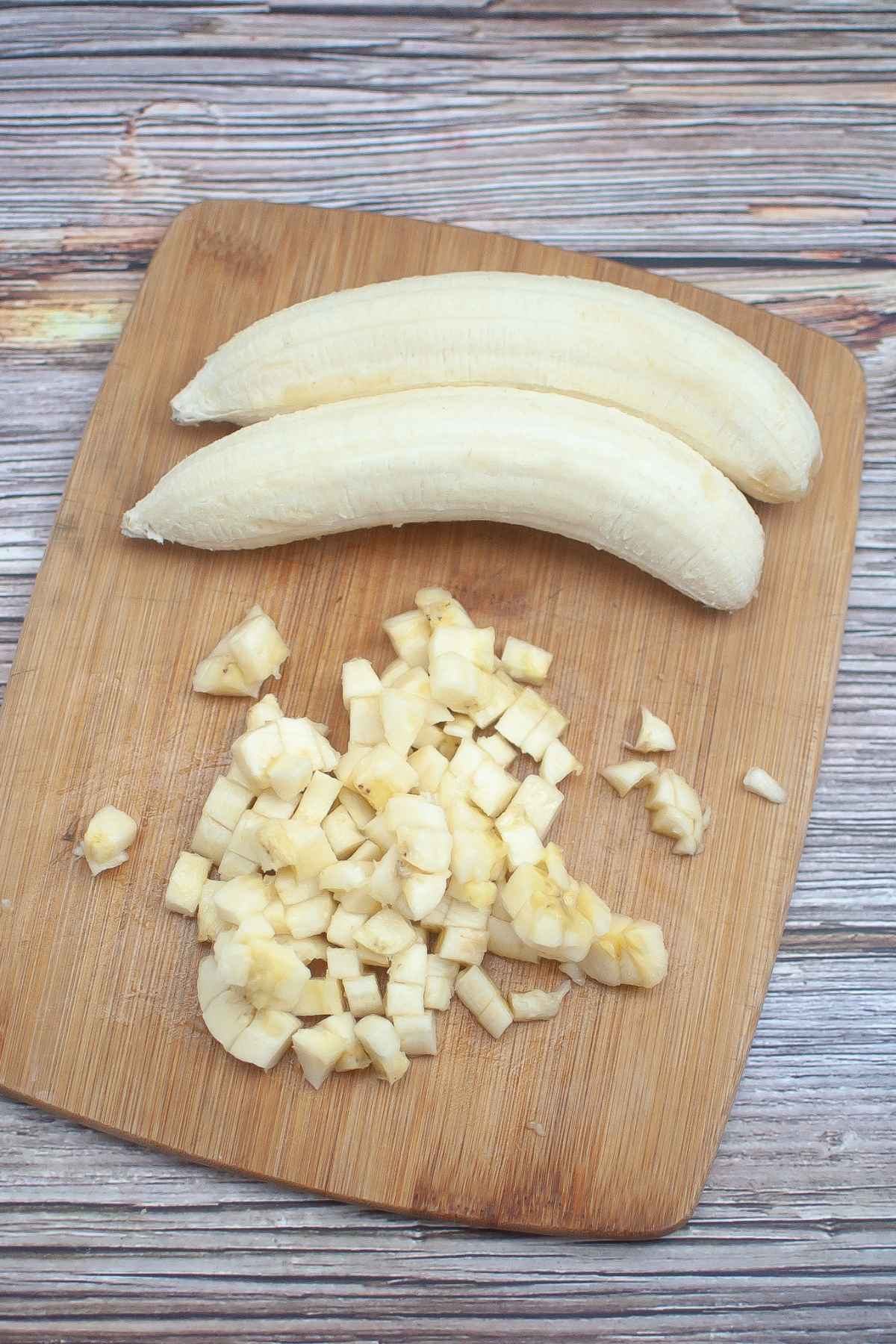 Two whole bananas on a cutting board with one diced banana in beneath them.