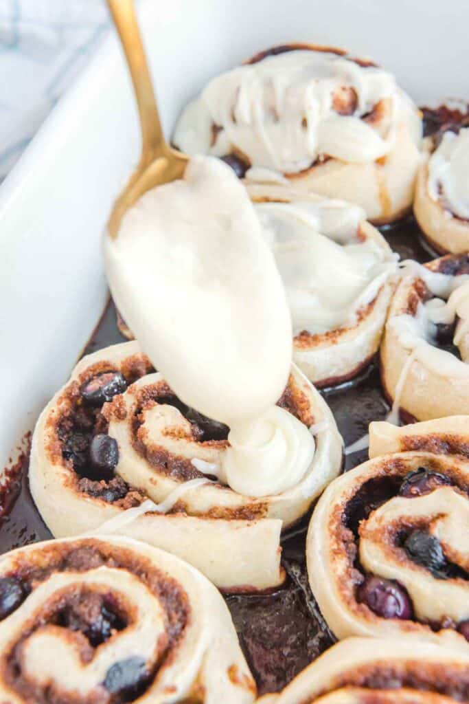 A gold spoon drizzling icing over top of baked cinnamon rolls.