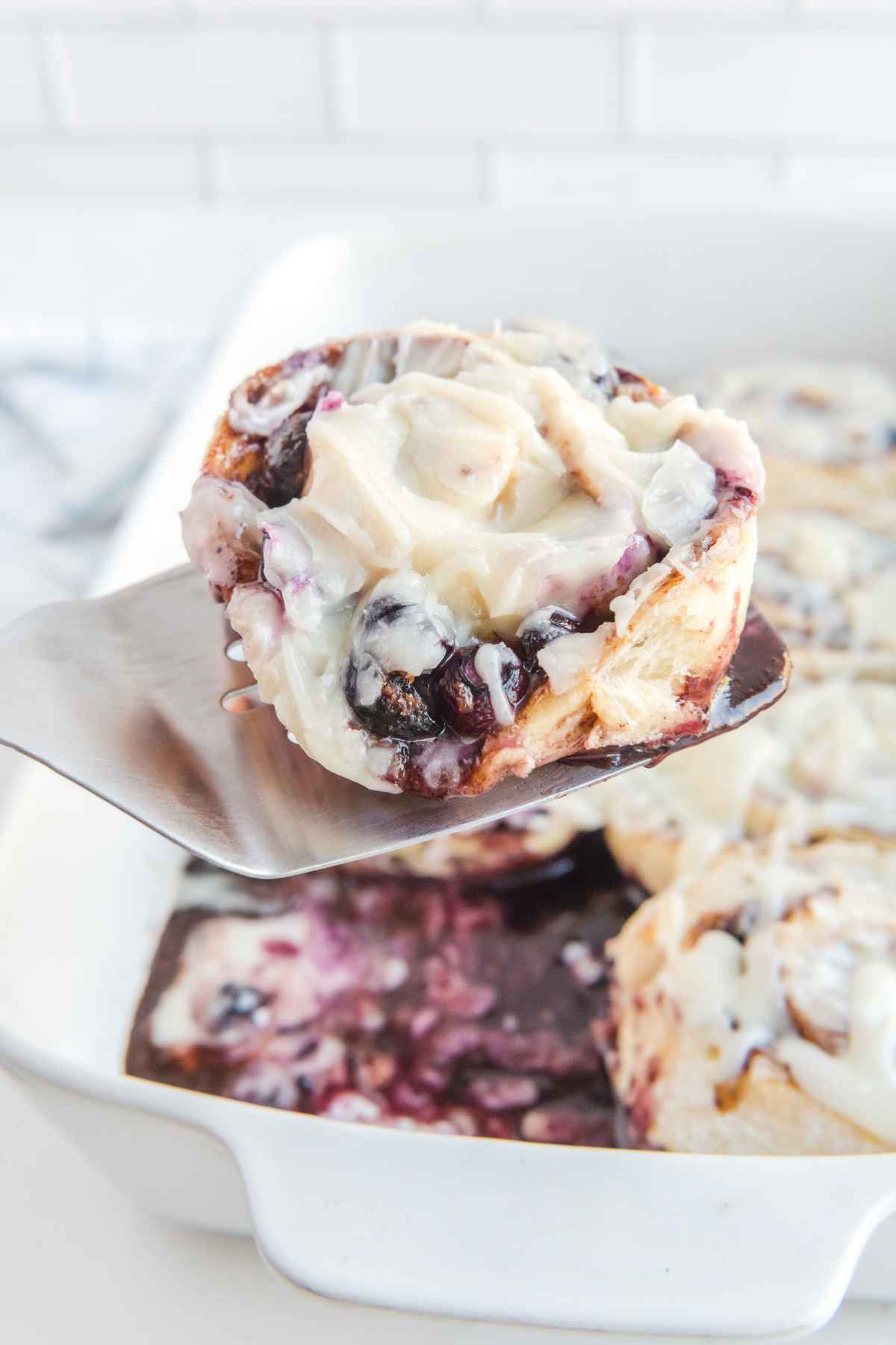 One blueberry cinnamon roll on a spatula hovering over the pan of rolls.