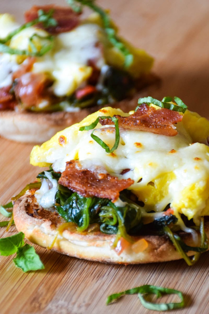 Breakfast Bruschetta made with a toasted english muffin topped with tomato spinach base, scrambled eggs, bacon, and cheese.