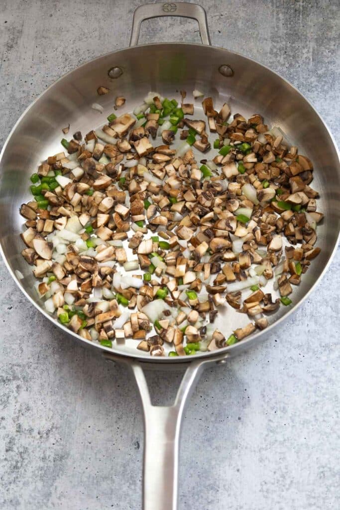 Mushrooms, peppers, and onions cooking in a stainless steel skillet.