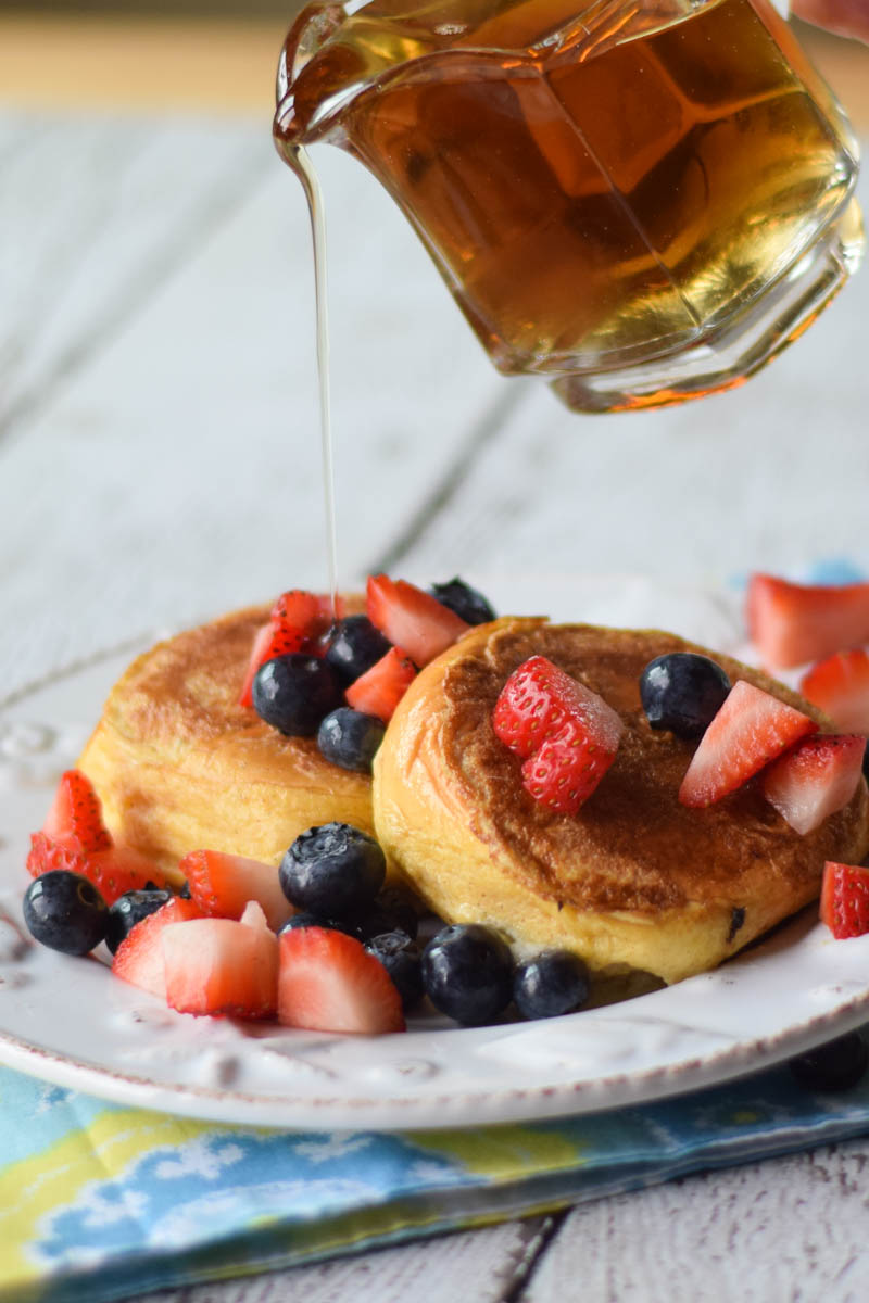 Pouring maple syrup over top of cheesecake stuffed french toast sliders topped with fresh blueberries and strawberries.