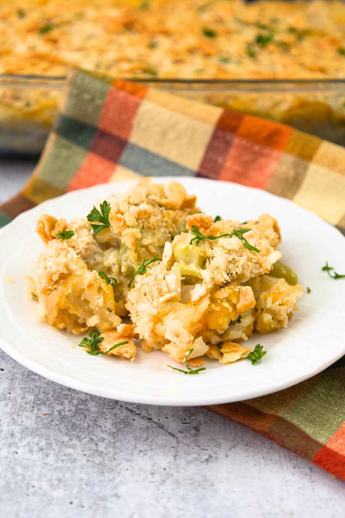 Side view of cheesy chicken broccoli rice casserole recipe on a white plate with blurred image of casserole dish in the background.