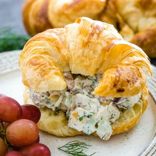 Chicken Salad on a croissant with red grapes next to the sandwichs and a piece of fresh dill in front of the sandwich with croissants blurred in the background.