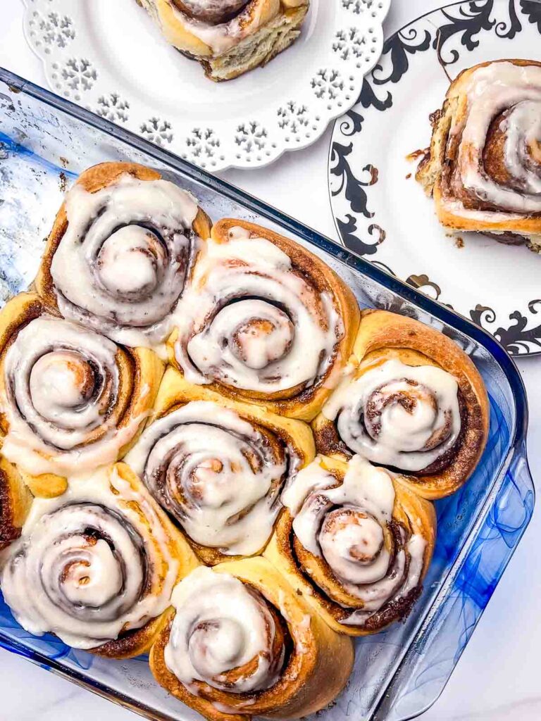 Overhead shot of frosted cinnamon rolls in a blue baking dish with tw rolls on plates to the upper right.