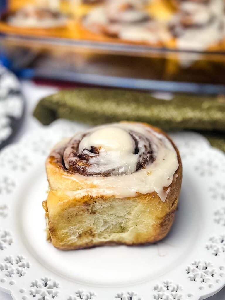 One clone of a cinnabon cinnamon roll on a white plate with the pan of rolls blurred in the background.