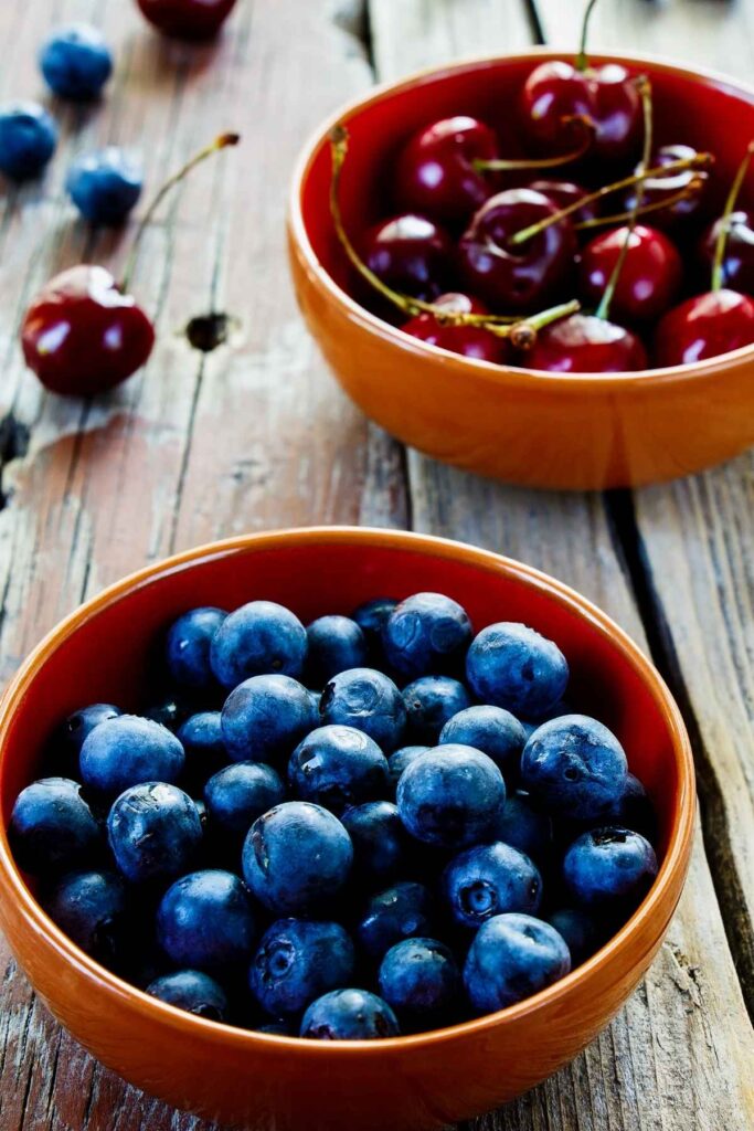 An orange bowl with blueberries and another orange bowl with stemmed cherries in the background.