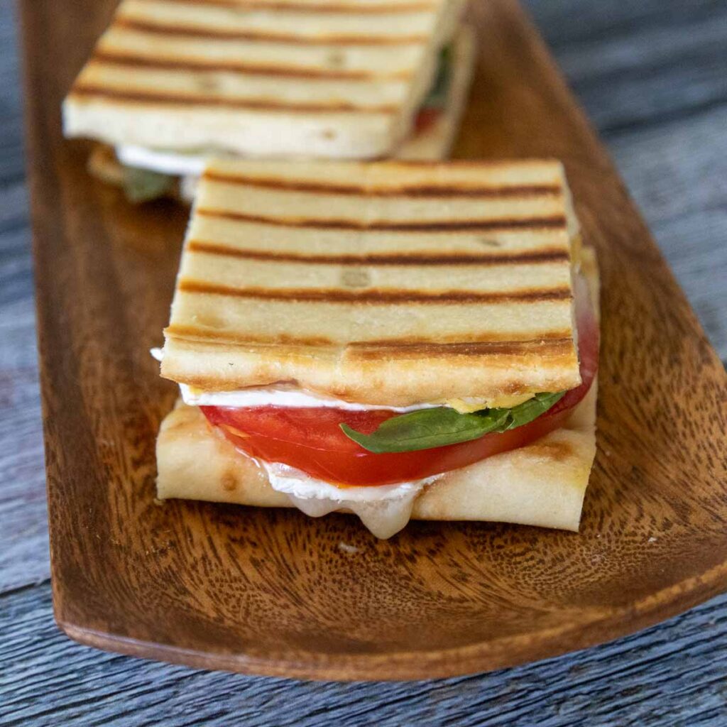 Two grilled brie tomato sandwichs made with flatbread with brie cheese, red tomato, and green fresh basil on a brown serving tray. 