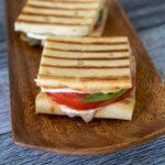 Two grilled brie tomato sandwichs made with flatbread with brie cheese, red tomato, and green fresh basil on a brown serving tray.