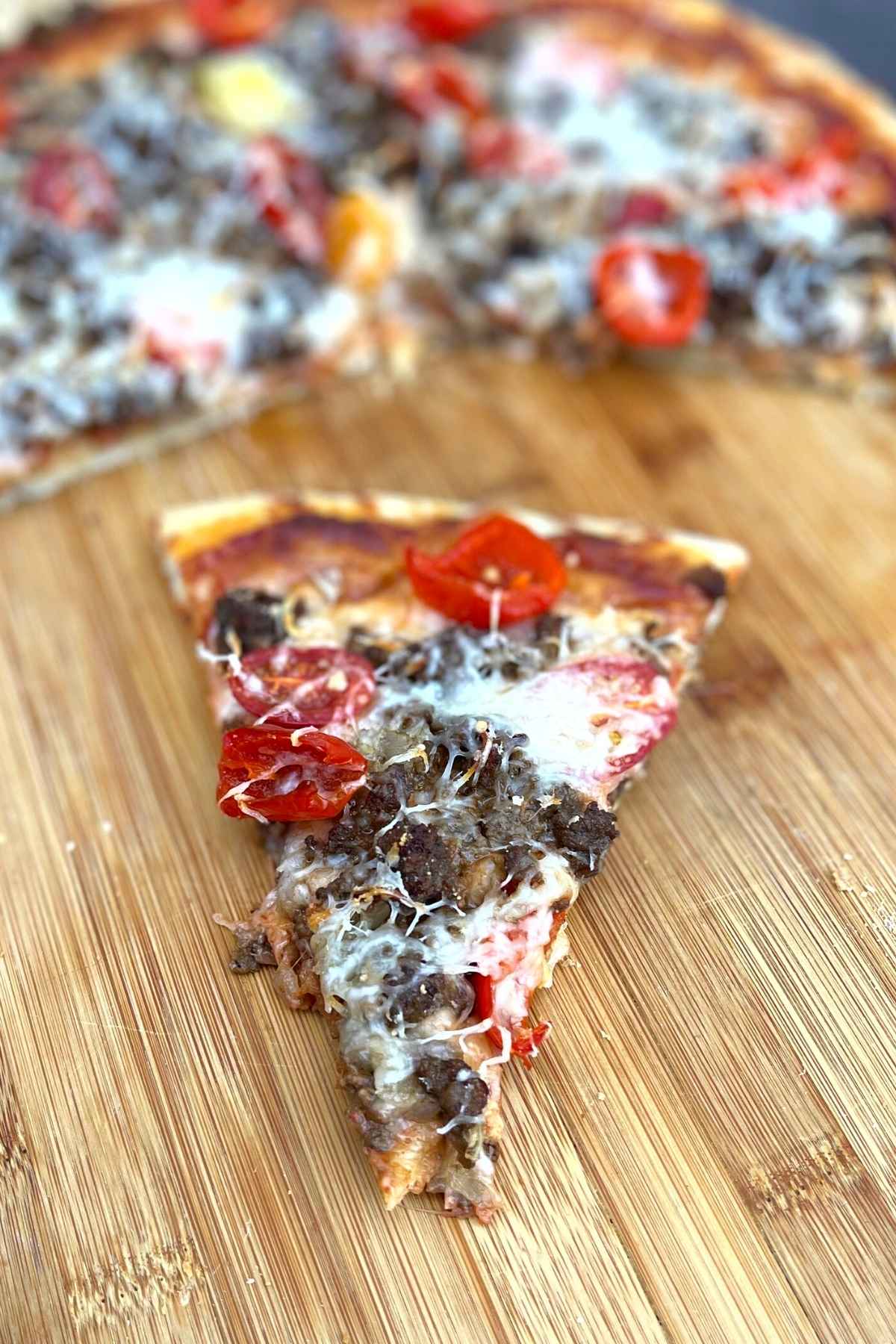 One slice of pizza facing point forward with whole pie blurred in the background.