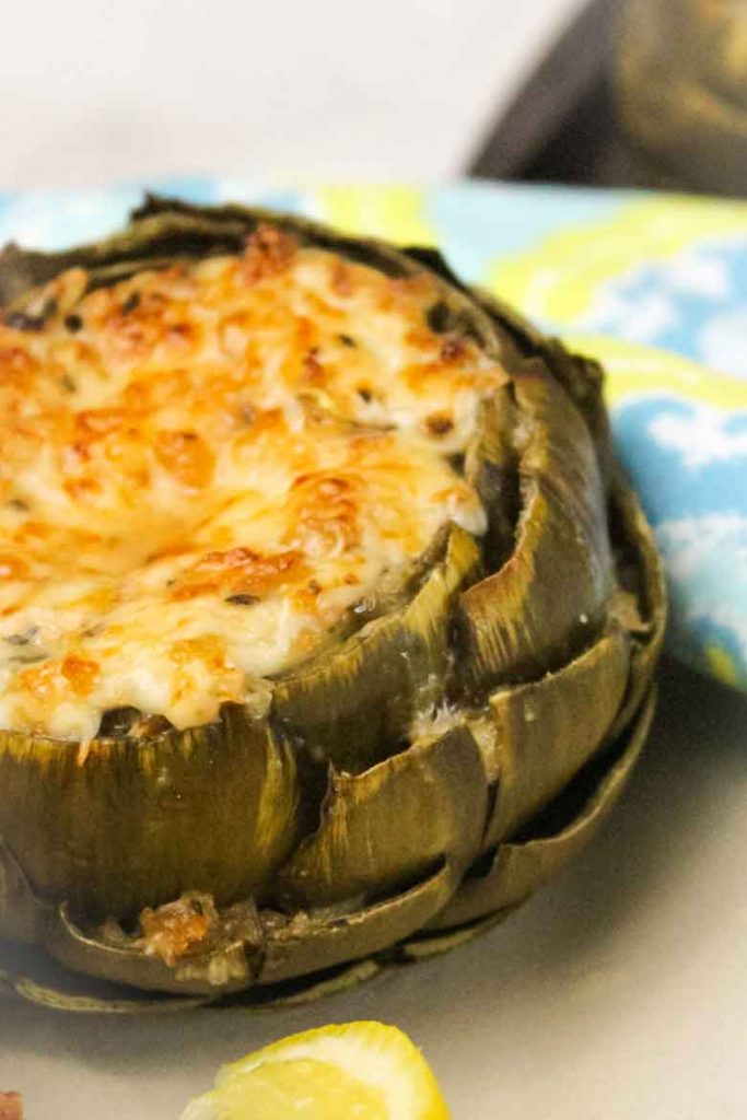 Cheese and Herb Stuffed Artichokes on a serving plate with a lemon slice and napkin in the background