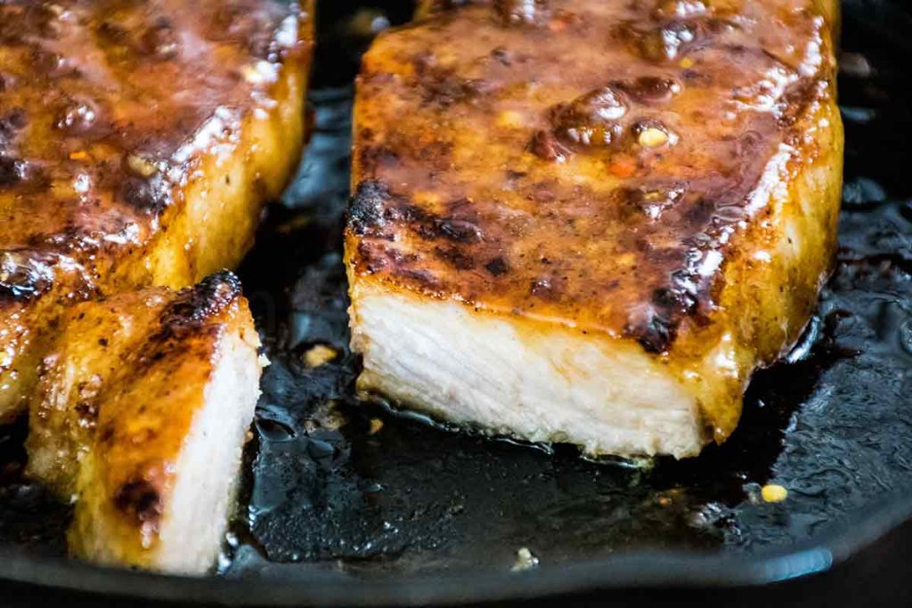 Two glazed Pork Chops in a cast-iron skillet with a piece of pork cut off to see the inside of the pork.