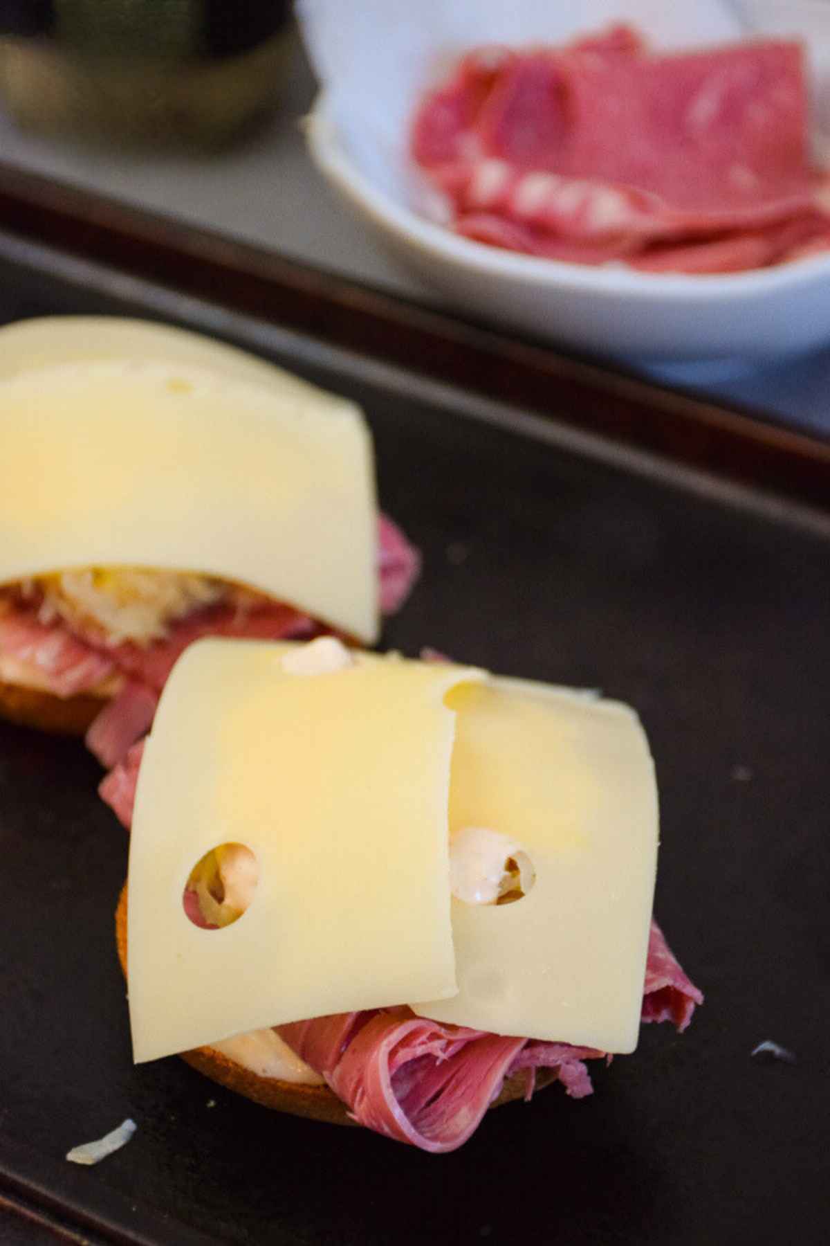 Two bagel halves topped with meat, sauce, sauerkraut and cheese on a baking sheet.