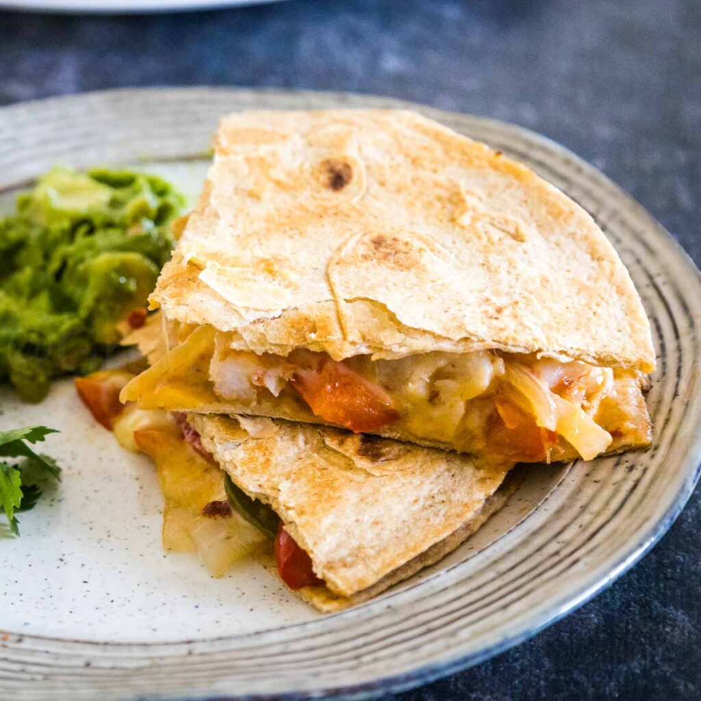 Toasty shrimp quesadillas on a cream colored plate cut into quarters with shrimp, red peppers, onions, and a side of guacamole in the background