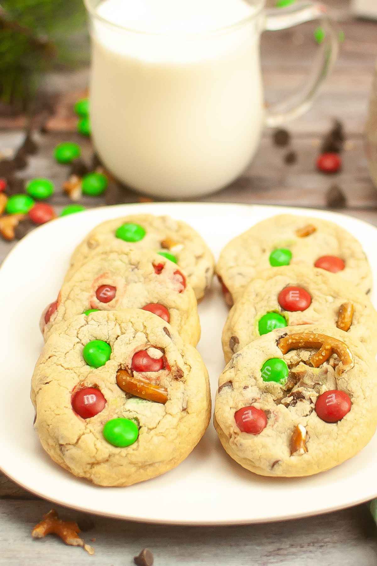 A white plate with six m&m pretzel cookies on it and a glass of milk in the background.