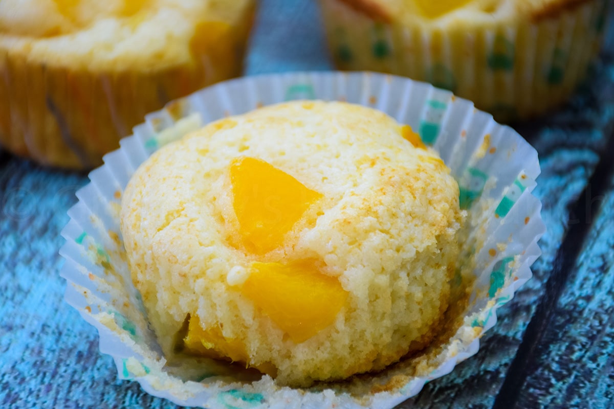 A baked muffin with peaches poking out of the top and paper wrapper removed.