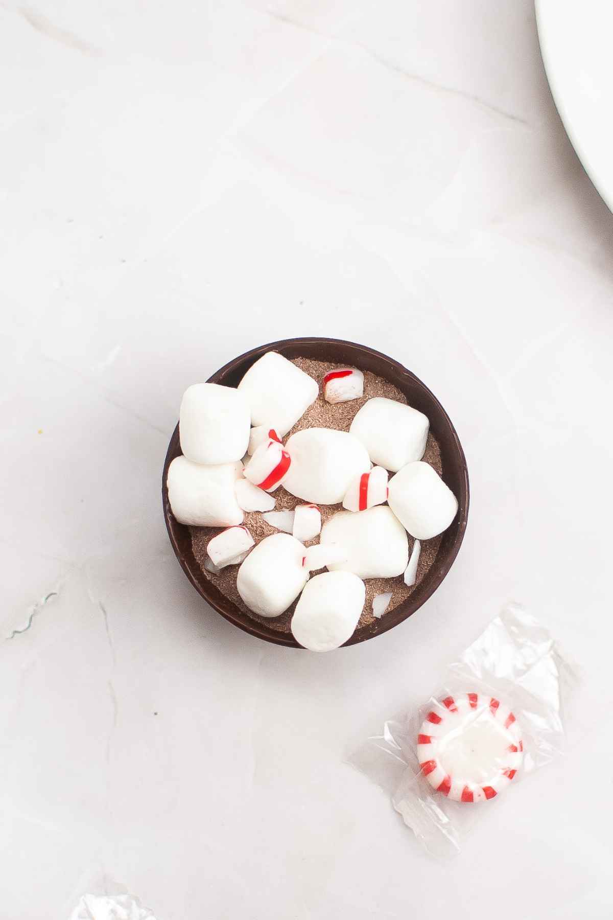 Overhead image of half chocolate shell filled with cocoa mix, mini marshmallows and crushed peppermint.