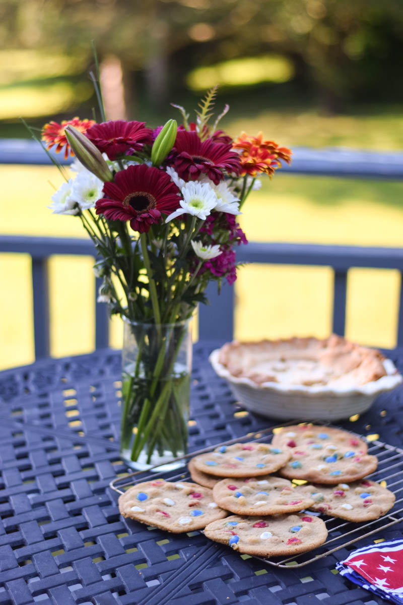 A wire rack on a table topped with cookies cooling, a bouquet of flowers and pie in the background