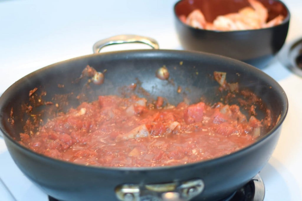 Tomato sauce in a skillet on the stove with a bowl of cooked shrimp blurred in the background.