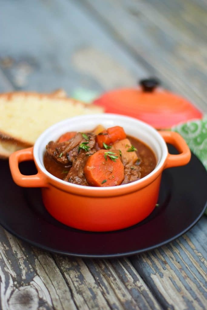 Slow cooked venison stew in an orange crock with slices of homemade bread in the background.