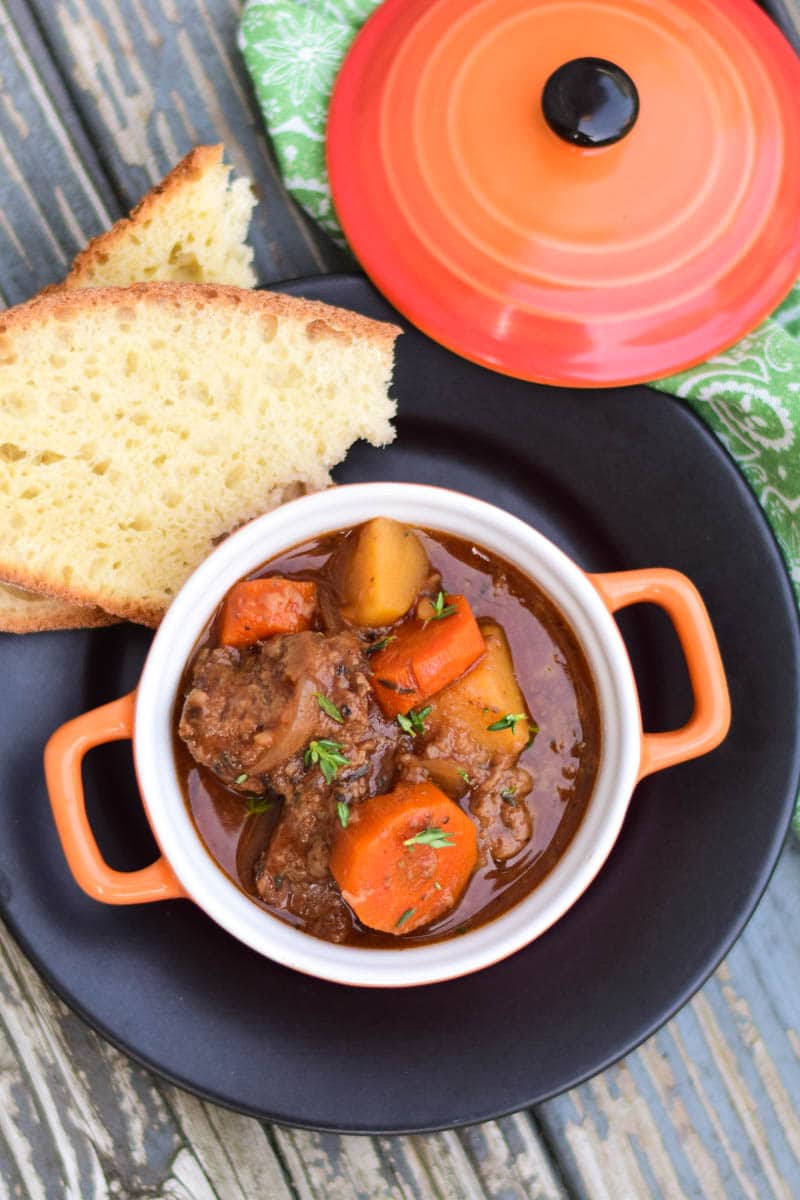 Crockpot Venison Stew in a orange serving crock sitting on a black plate with two slices of homemade bread.