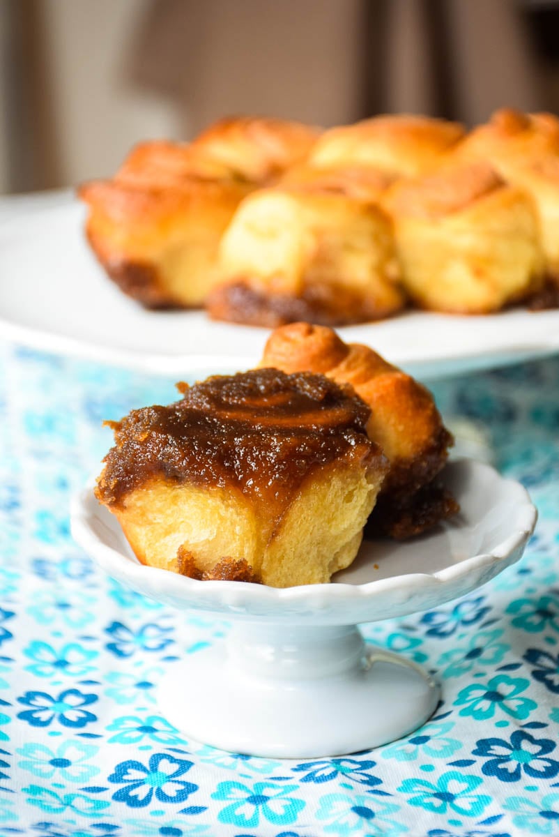 One serving crescent roll sticky buns on a plate with a large plate of buns blurred in the background.