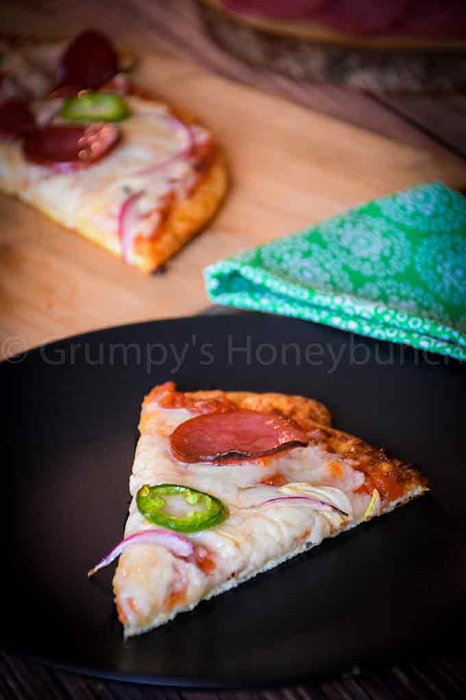 A slice of salami pizza on a black plate with more pizza in the background