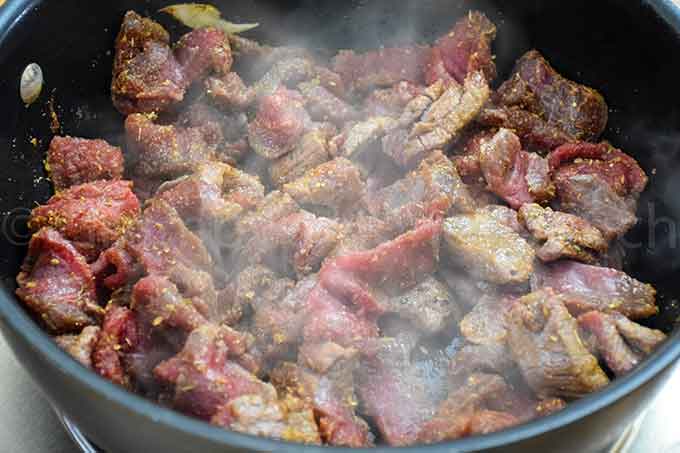 Partially cooked strips of seasoned steak in a large skillet with steam rising.