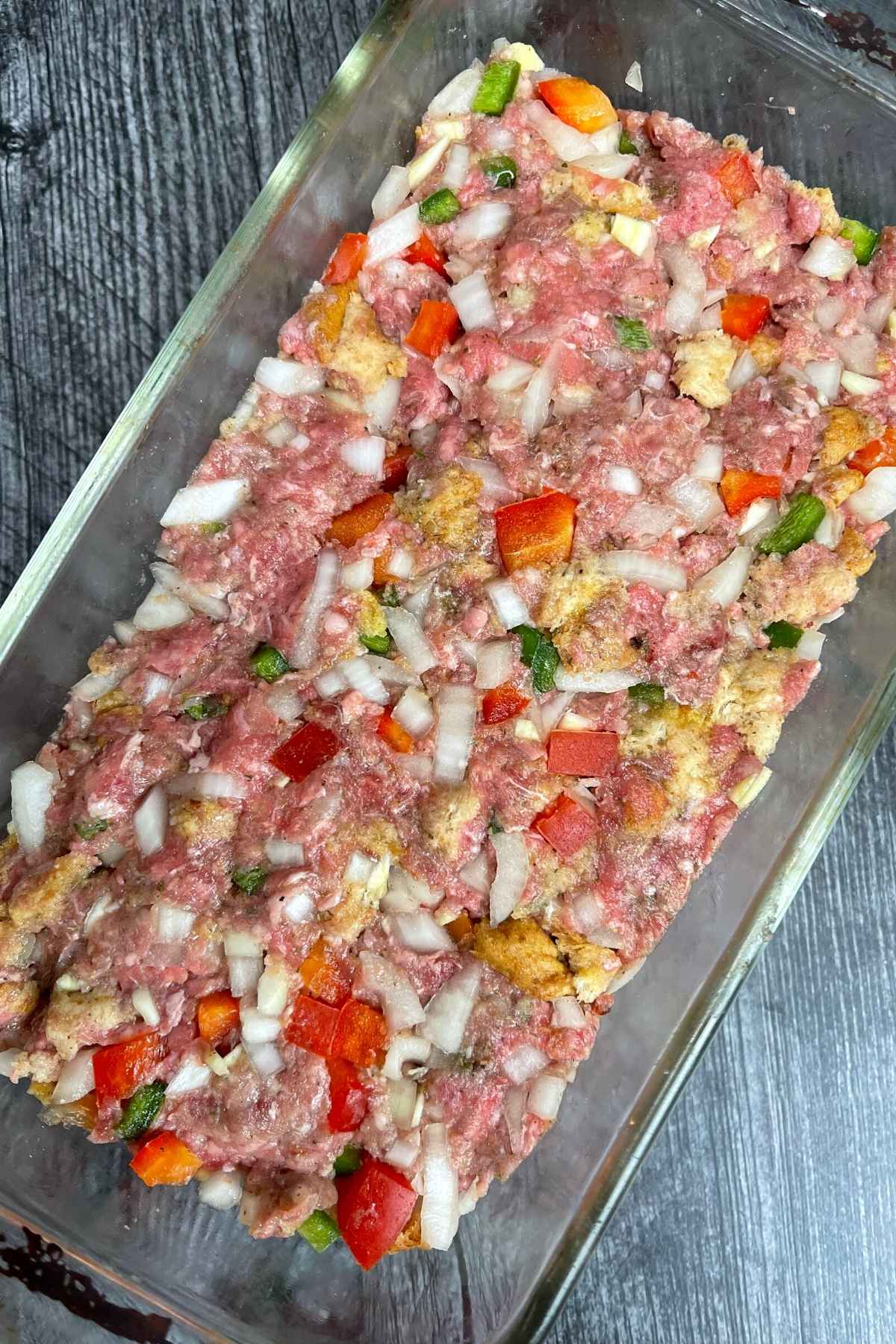 Raw meatloaf in a baking dish with specks of bread crumbs, peppers, and onions visible.