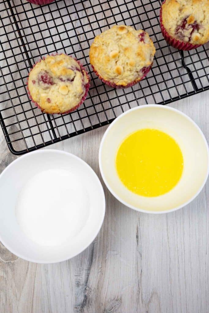 A bowl of white sugar and bowl of melted butter with baked muffins on a cooling rack.