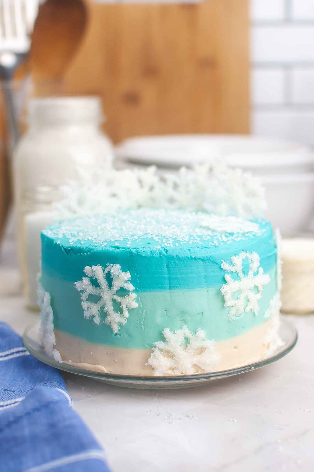 Side view of a layer cake with blue ombre frosting, and sugar glitter white chocolate frosting decorations.
